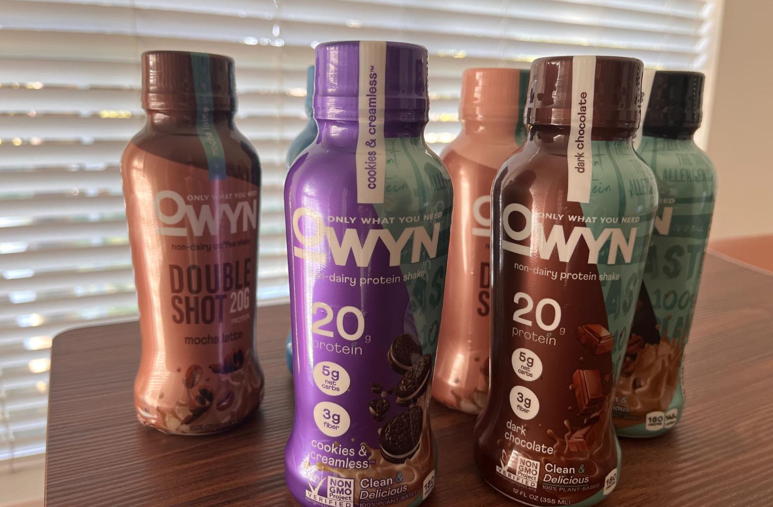 Five bottles OWYN Plant-Based Protein Shakes on a kitchen table, including the flavors Cookies & Creamless, Dark Chocolate and Double Shot Mocha Fudge